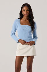 A woman wearing a light blue, long-sleeved, ribbed sweater with a square neckline and scalloped edges, paired with a high-waisted, white mini skirt.
